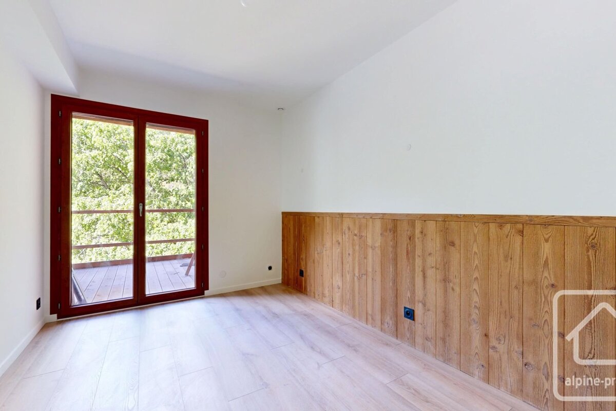 An empty, clean room with white walls and light wood flooring. An open wooden door reveals another room, and a white heater is visible on the left wall.