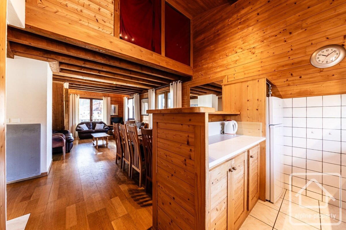 A rustic wooden chalet interior with open-plan kitchen, dining area, and living room with leather sofas. A mezzanine level with red curtains is visible.