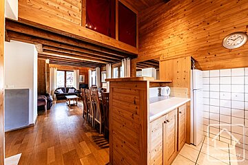 A rustic wooden chalet interior with open-plan kitchen, dining area, and living room with leather sofas. A mezzanine level with red curtains is visible.