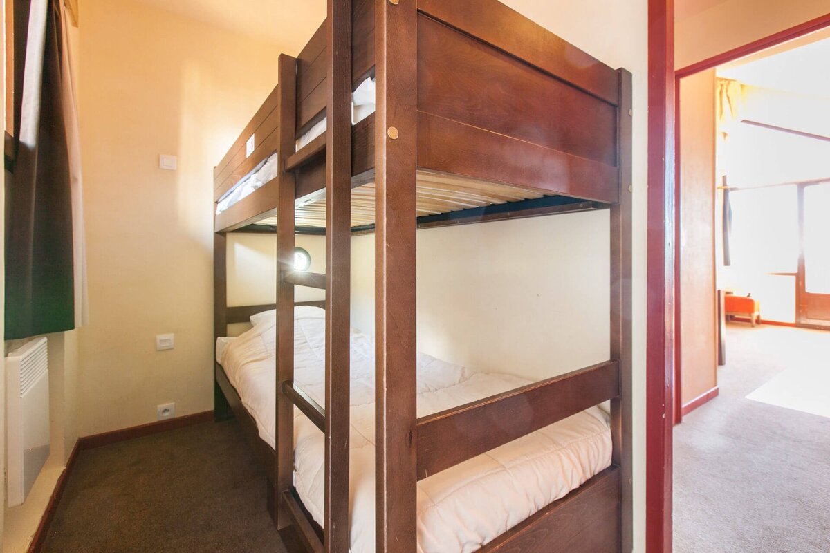 A cozy room featuring wooden bunk beds with white bedding, dark carpet, and light walls. A doorway on the right leads to a brightly lit area.