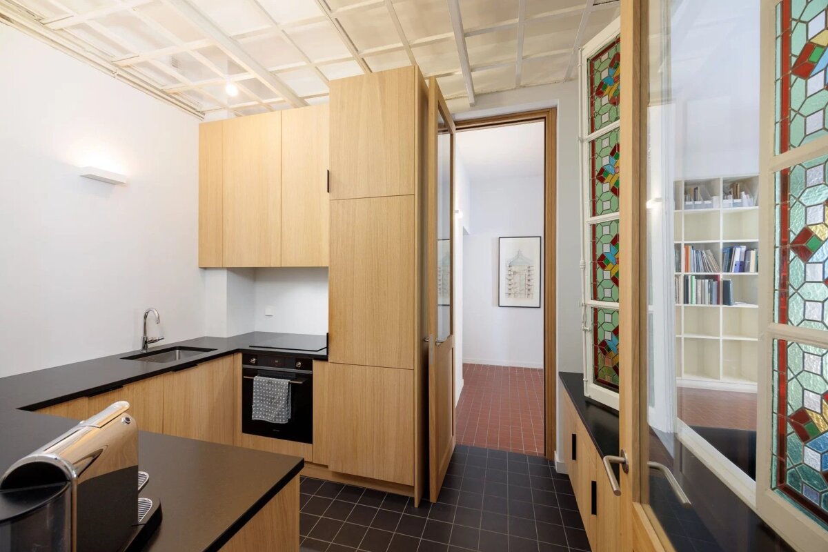 Modern kitchen featuring light wood cabinetry, black countertops, and a stained glass window partition beside a doorway.