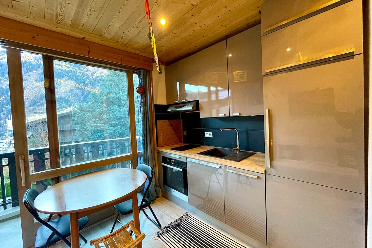 A modern kitchenette with glossy grey cabinets and a small dining table next to a large window overlooking a mountainous, forested landscape.