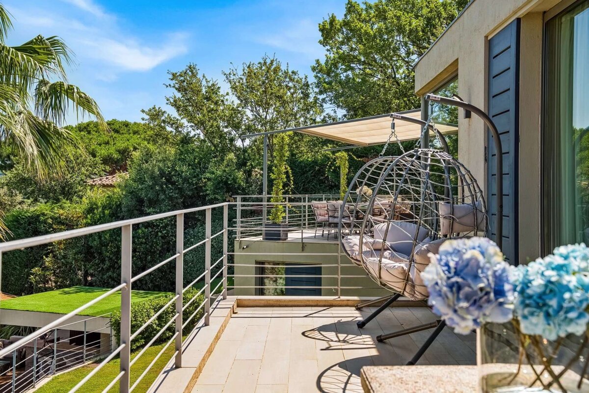 A modern, sunlit balcony features a hanging egg chair, blue hydrangeas, and views of lush green trees under a clear blue sky.