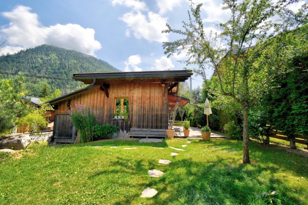 Rustic wooden cabin with a stone path on a green lawn, set against a backdrop of lush, forested mountains.