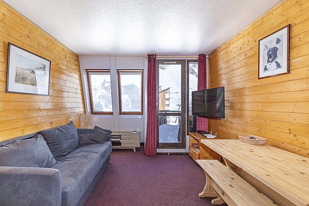 A cozy, wood-paneled room with a gray sofa, wooden dining table, TV, and balcony door looking out onto a snowy mountain landscape.