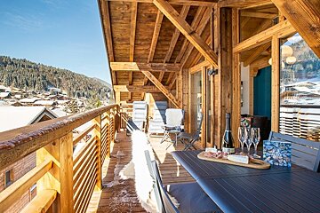 Wooden chalet balcony featuring a table with wine and charcuterie, overlooking a snowy alpine mountain village.