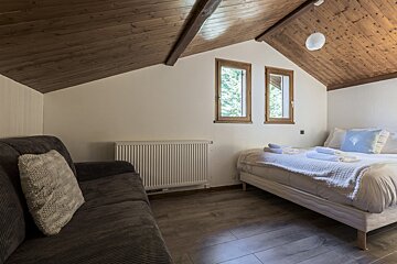 Cozy attic room with a sloped wooden ceiling, a bed, a sofa, and two windows offering a glimpse of nature. Hardwood floors and a radiator complete the space.