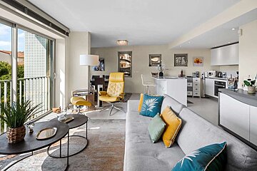 Bright, modern open-plan living room and kitchen with a balcony view, gray sofa, yellow armchair, and colorful accents.