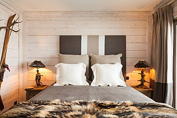 A cozy, rustic chalet bedroom. Light wood walls, a large bed with neutral bedding, a faux fur throw, and unique lamps. Skis & antlers hang on the wall.