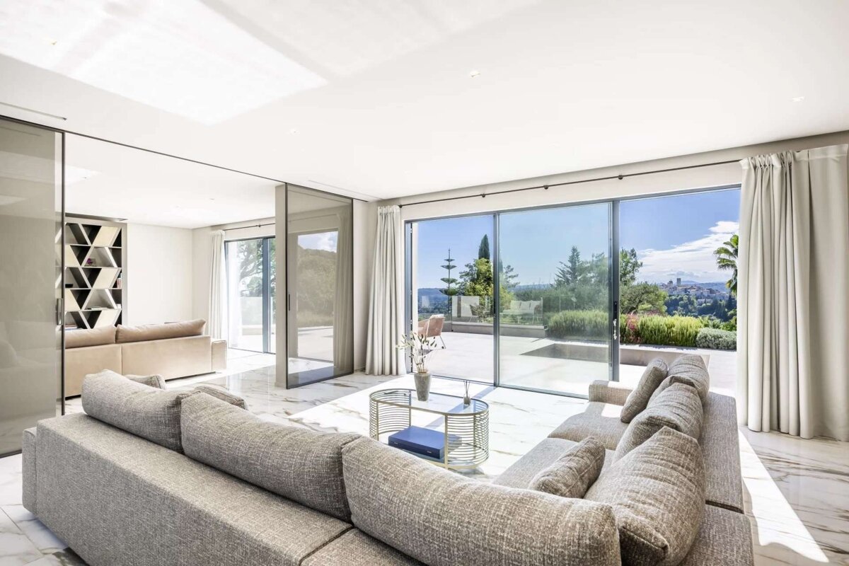 A bright, modern living room with a large grey sectional sofa, marble floors, and sliding glass doors revealing a sunny panoramic view of a lush landscape and distant town.