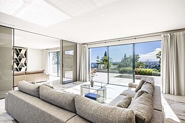 A bright, modern living room with a large grey sectional sofa, marble floors, and sliding glass doors revealing a sunny panoramic view of a lush landscape and distant town.