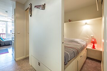 A compact home interior with a hallway leading to a living room, and a built-in bed nook with a red lamp and patterned duvet on the right.