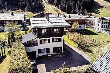 Aerial view of chalet-style houses nestled in a green mountain valley with pine forests. The main house features dark wood and white walls; a car is parked nearby.
