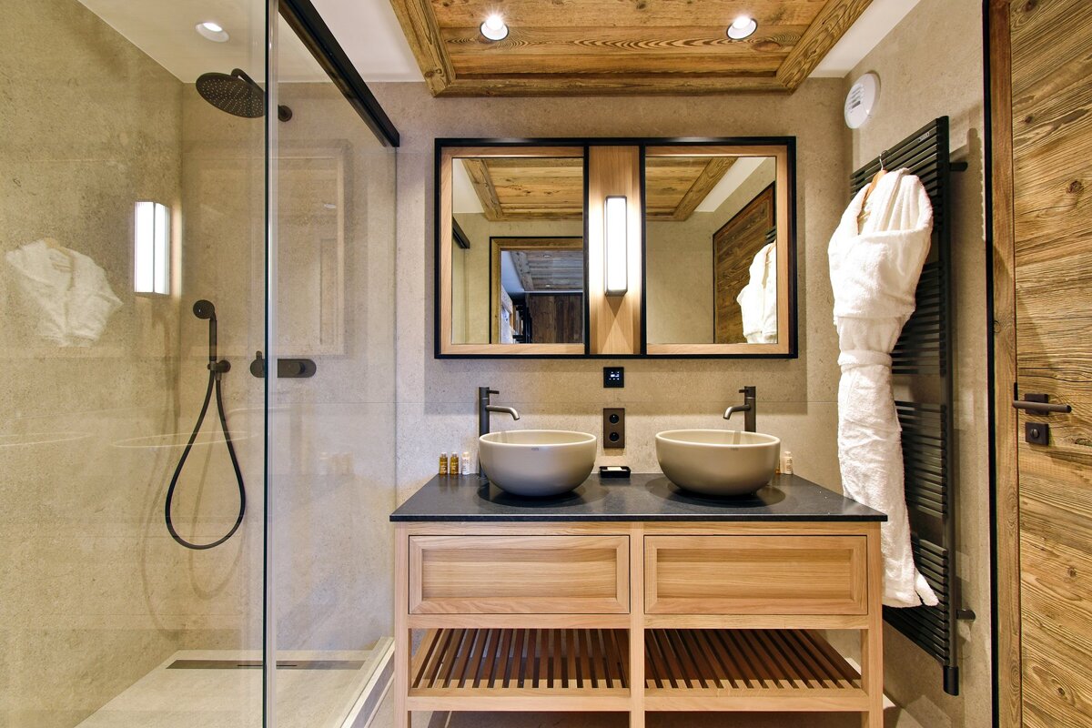 An elegant bathroom with a double wooden vanity, vessel sinks, modern mirrors, and a glass shower. Rustic wooden ceiling and door complement the spa-like decor.