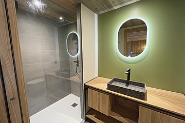 A modern bathroom features a glass shower next to a vanity with a speckled sink, black faucet, and oval backlit mirror on a green wall.