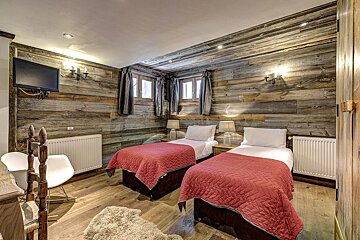 Rustic bedroom with two beds, red quilts, wood paneling on walls/ceiling, small windows, a TV, and a cozy, inviting atmosphere.
