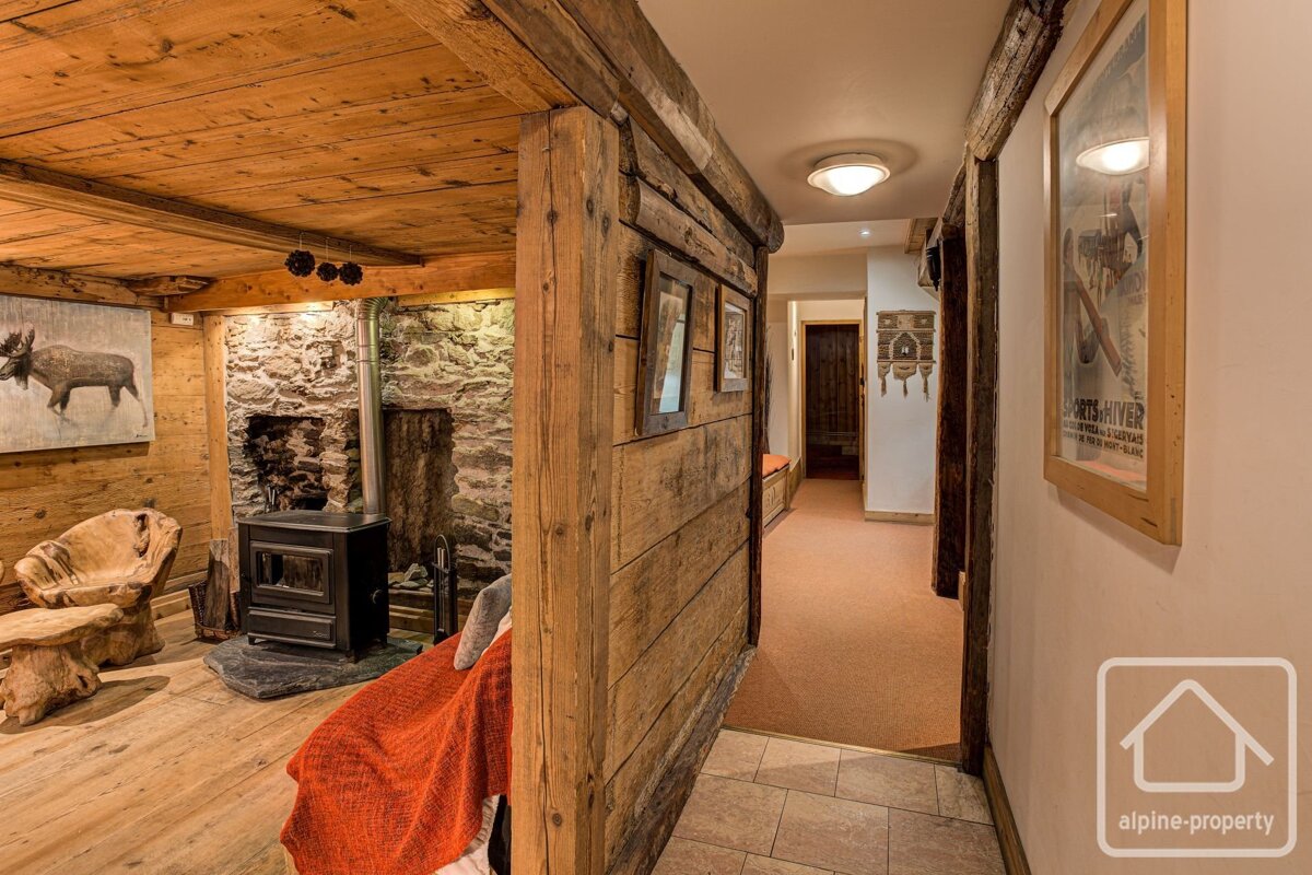 A rustic, chalet-style interior featuring wood paneling, a stone fireplace with a wood stove, and a carpeted hallway with framed art.