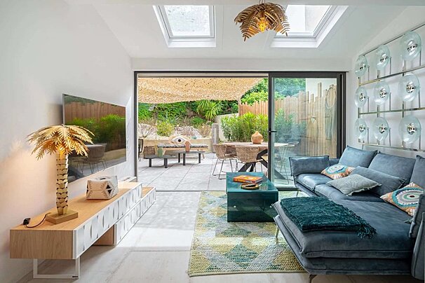 Modern living room with a blue sectional sofa, a gold palm tree lamp, and skylights. Large glass doors open to a lush patio with dining and lounge areas.