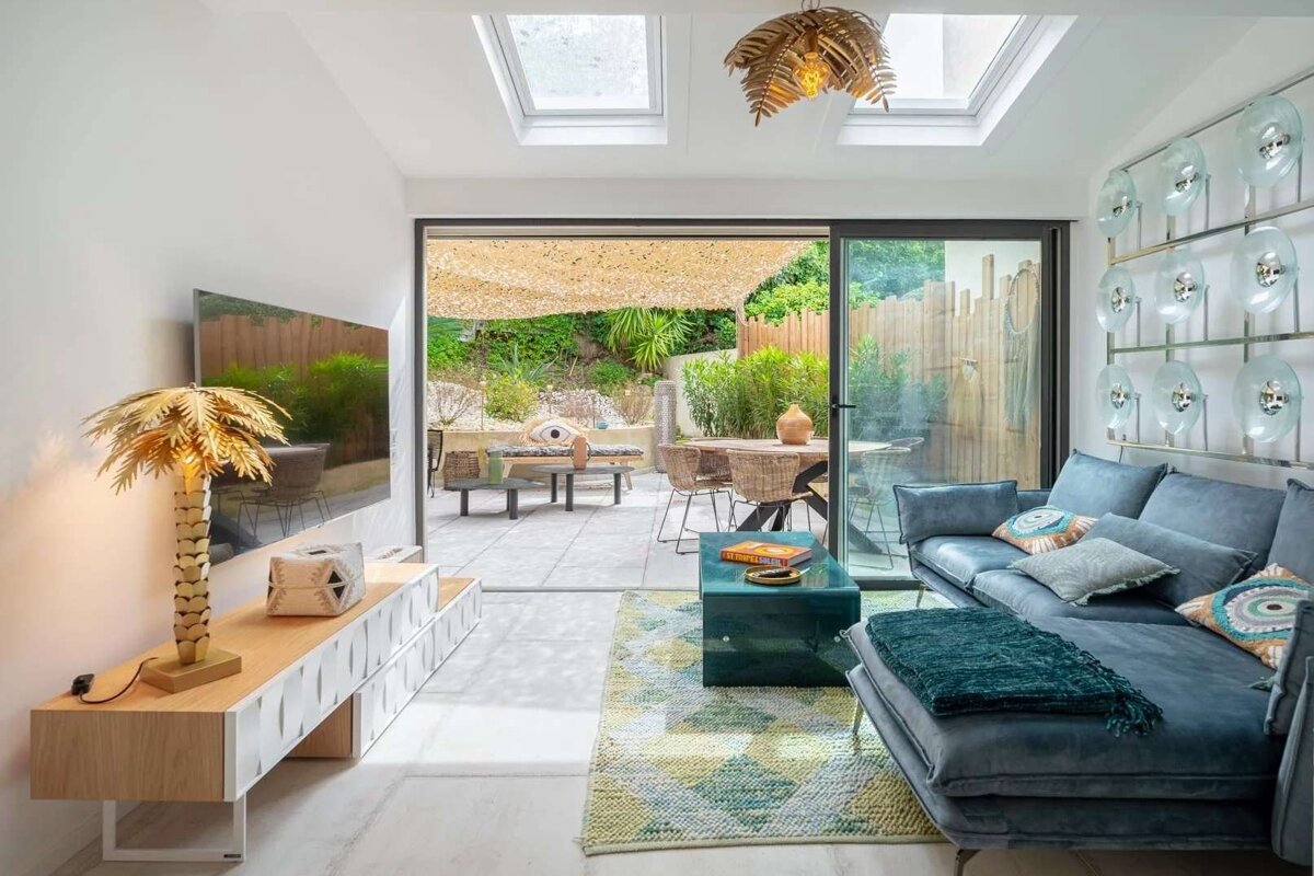 A chic, modern living room with a blue sectional sofa, a palm tree lamp, and large glass doors opening to a lush patio with outdoor seating. Skylights brighten the space.