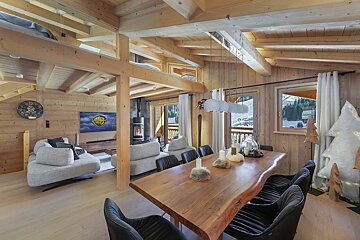 Cozy, rustic wooden chalet interior with open-plan living and dining, featuring a live-edge table, fireplace, comfy sofas, and snowy mountain views.