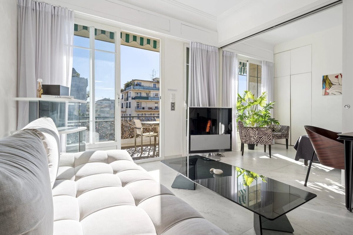 A bright, modern white living room with glass furniture and open doors leading to a balcony.