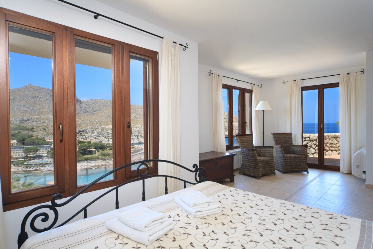 Bright bedroom with large windows, showcasing scenic views of mountains, a bay, and the sea. Features a bed and wicker chairs.