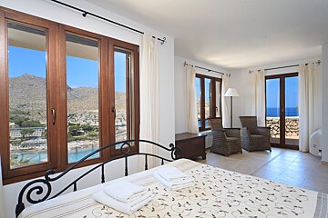 Bright bedroom with large windows, showcasing scenic views of mountains, a bay, and the sea. Features a bed and wicker chairs.