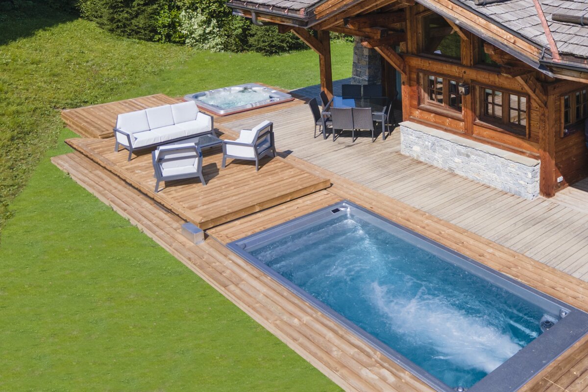 An aerial view of a luxurious chalet with a wooden deck, swimming pool, hot tub, and outdoor seating, surrounded by green grass on a sunny day.