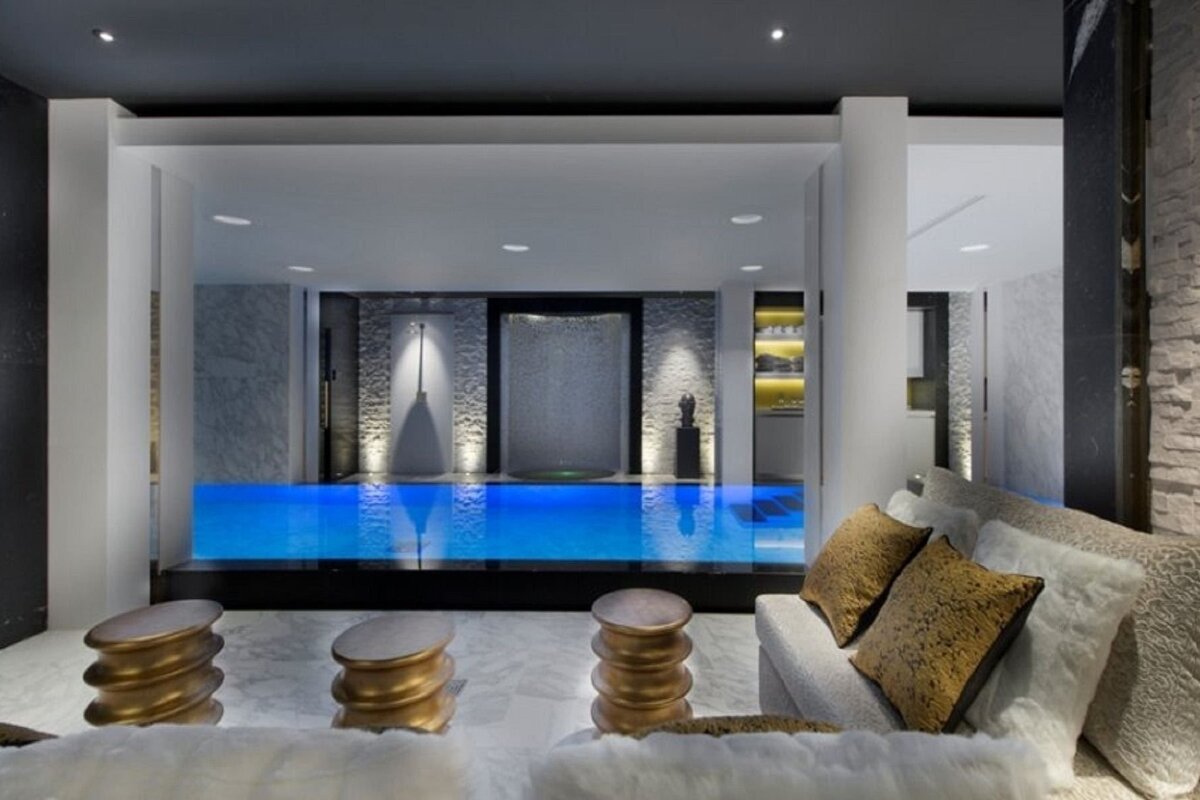 A modern indoor living space featuring a glowing blue pool, an elegant water feature, and a stylish seating area with golden stools.
