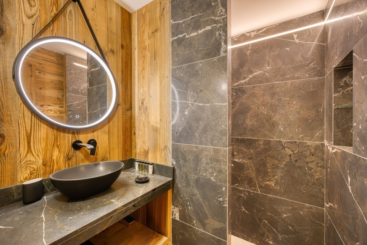 Modern bathroom with a lighted round mirror, black vessel sink, wooden accent wall, and dark marble tiles.