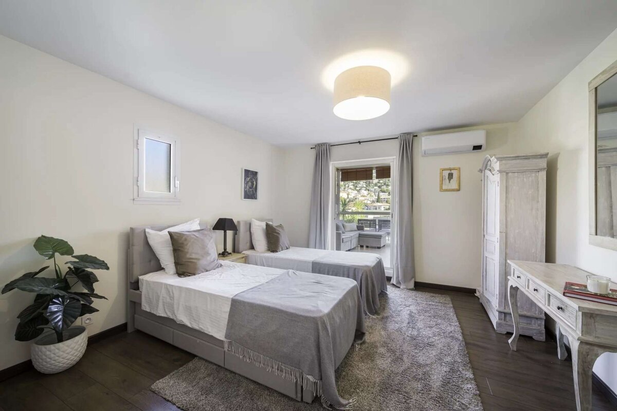 A bright, minimalist twin bedroom with two single beds, light grey and white decor, a large window, and a balcony view.
