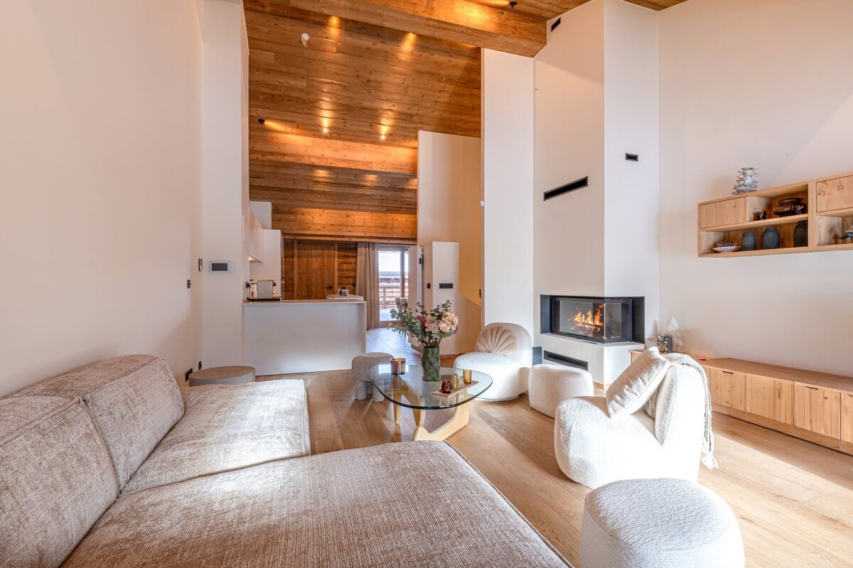 Bright modern chalet living room with a sectional sofa, white chairs, glass coffee table, fireplace, and high wood-beamed ceiling.