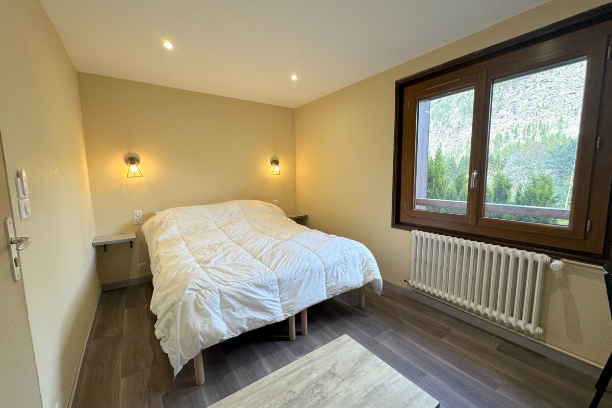 A simple bedroom with yellow walls, a white bed, wooden floor, and a large window showcasing a mountain forest view.