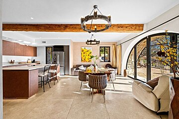 Open-plan living space with a modern kitchen island, dining table, sofa, rustic wood beam, ornate chandeliers, and arched glass doors overlooking an outdoor area.