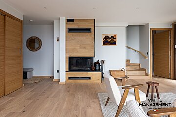 A modern, light-filled living room with wood floors, a prominent wood-paneled fireplace, mid-century armchair, and a staircase.