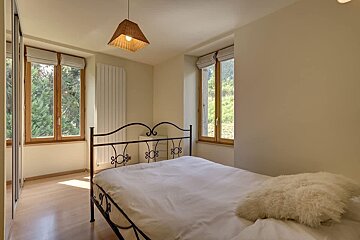 A cozy bedroom with a wrought-iron bed, white bedding, wooden floors, and two windows showcasing lush green nature outside.