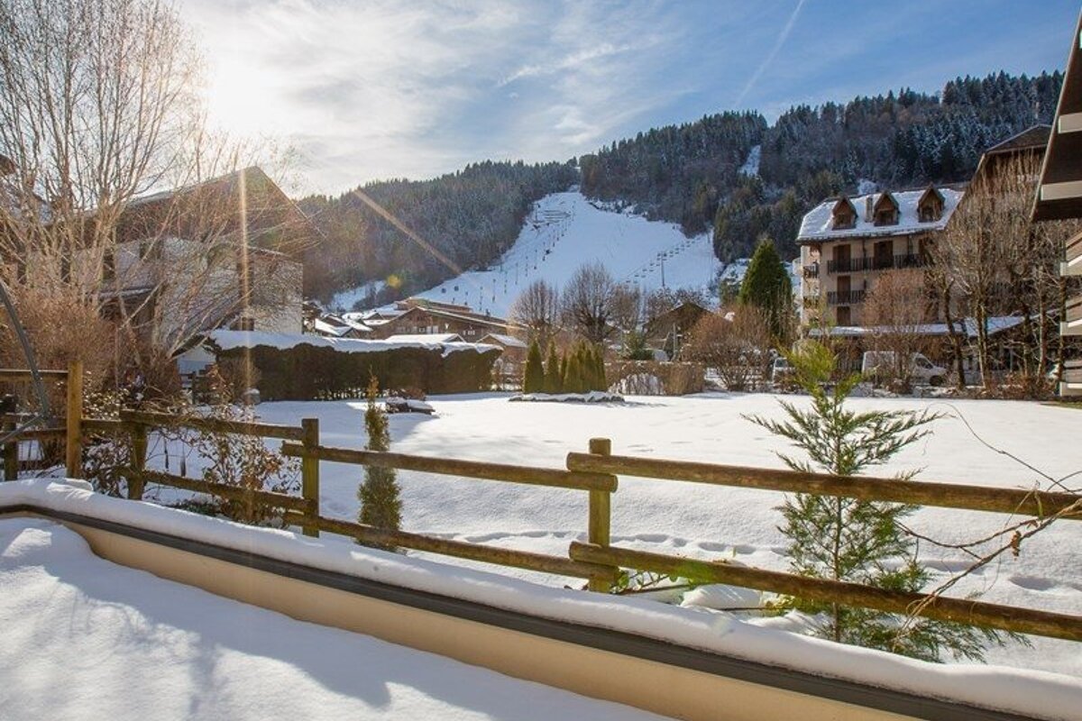 Sunny winter view of a snowy mountain village with chalets, a wooden fence, and a ski slope in the background.
