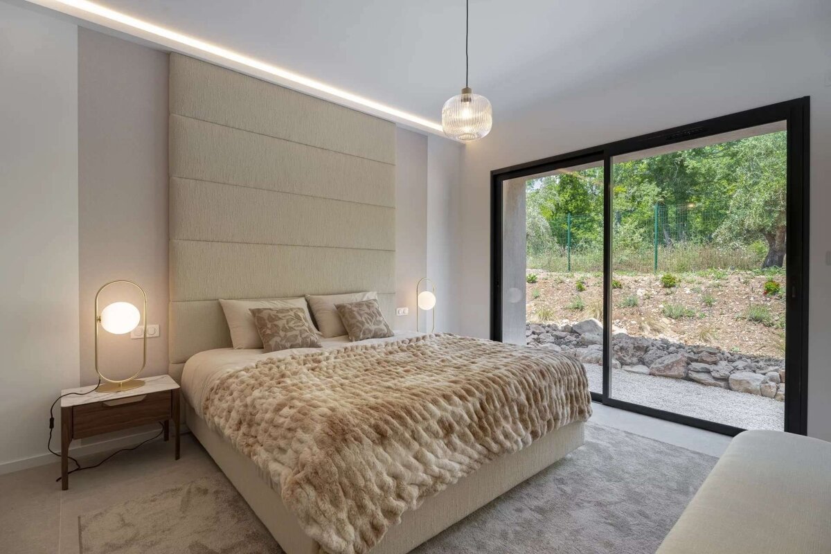 A modern, neutral-toned bedroom with a large bed, plush blanket, and a sliding glass door offering a view of a green outdoor landscape.