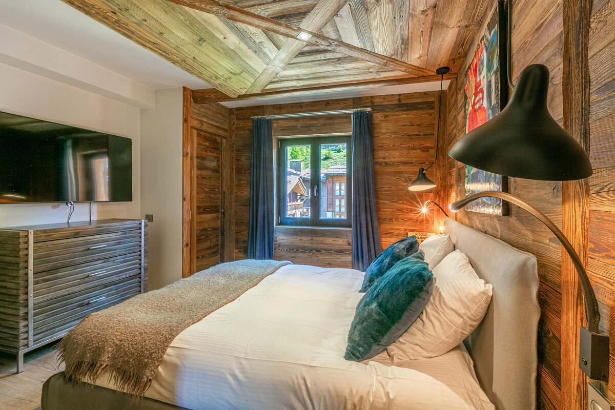 A modern chalet bedroom with warm wood paneling on walls/ceiling, a comfy bed, window view, TV, and stylish lamps for a cozy ambiance.