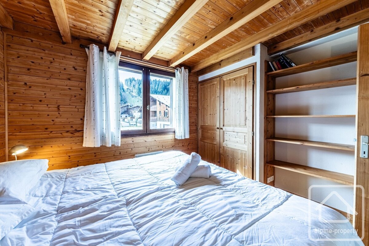 A rustic wooden bedroom features a large bed, a window with a view of a snowy village and mountains, a wardrobe, and a bookshelf.