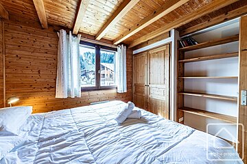 A rustic wooden bedroom features a large bed, a window with a view of a snowy village and mountains, a wardrobe, and a bookshelf.
