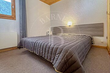 A simple bedroom features a bed with a grey comforter, a wooden headboard, a wall lamp, a window with a grey curtain, and a wooden ceiling.