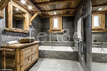 A modern chalet bathroom with rustic wooden accents, a stone sink, dark grey tiled walls, and a built-in bathtub.