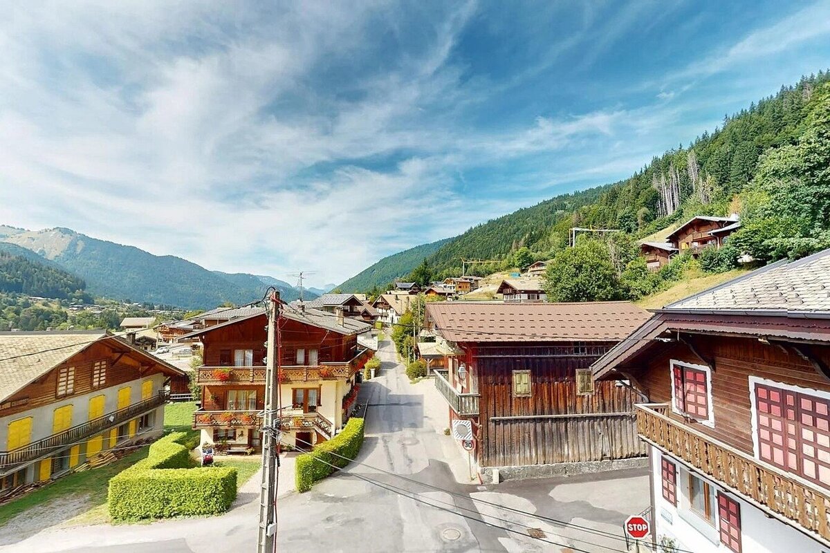 A picturesque mountain village with traditional wooden houses lining a street, framed by green mountains under a bright blue sky.