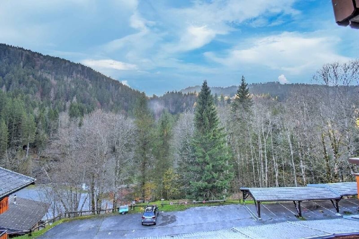 A mountain landscape with a parking lot and covered structure, flanked by evergreen and bare trees under a partly cloudy sky.