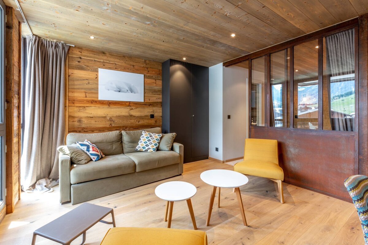 Cozy, rustic living room featuring wood-paneled walls/ceiling, a beige sofa, yellow chair, and a distinctive glass partition.