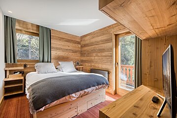 Cozy, rustic bedroom with warm wood-paneled walls, a comfortable bed, a modern TV, and a balcony door overlooking lush greenery.