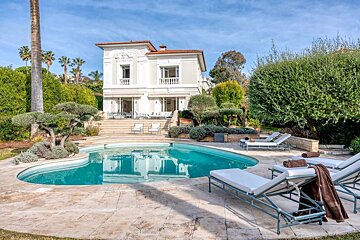 Elegant white villa with a beautiful curved swimming pool, surrounded by lush green landscaping and palm trees under a sunny blue sky.
