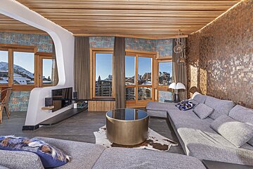 Modern living room with ski resort views, a grey sectional, gold coffee table, wooden ceilings, and unique textured walls.