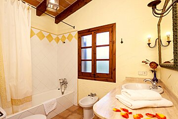 A warm, rustic bathroom with a tub, wooden window, bidet, and sink. Yellow walls, tiled accents, wooden beams, and rose petals create a cozy ambiance.